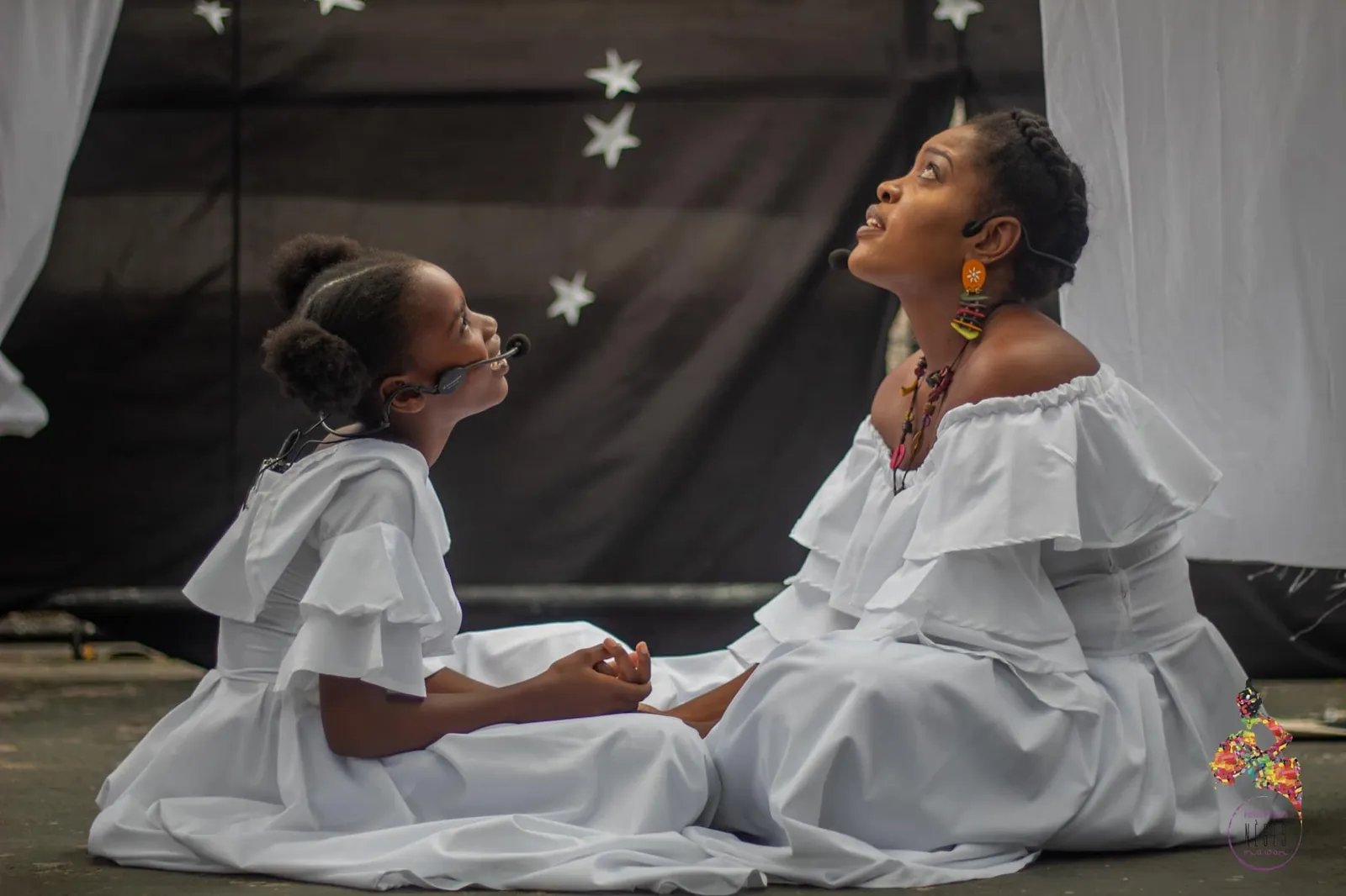 A girl and a woman in white ruffled dresses sitting on a stage