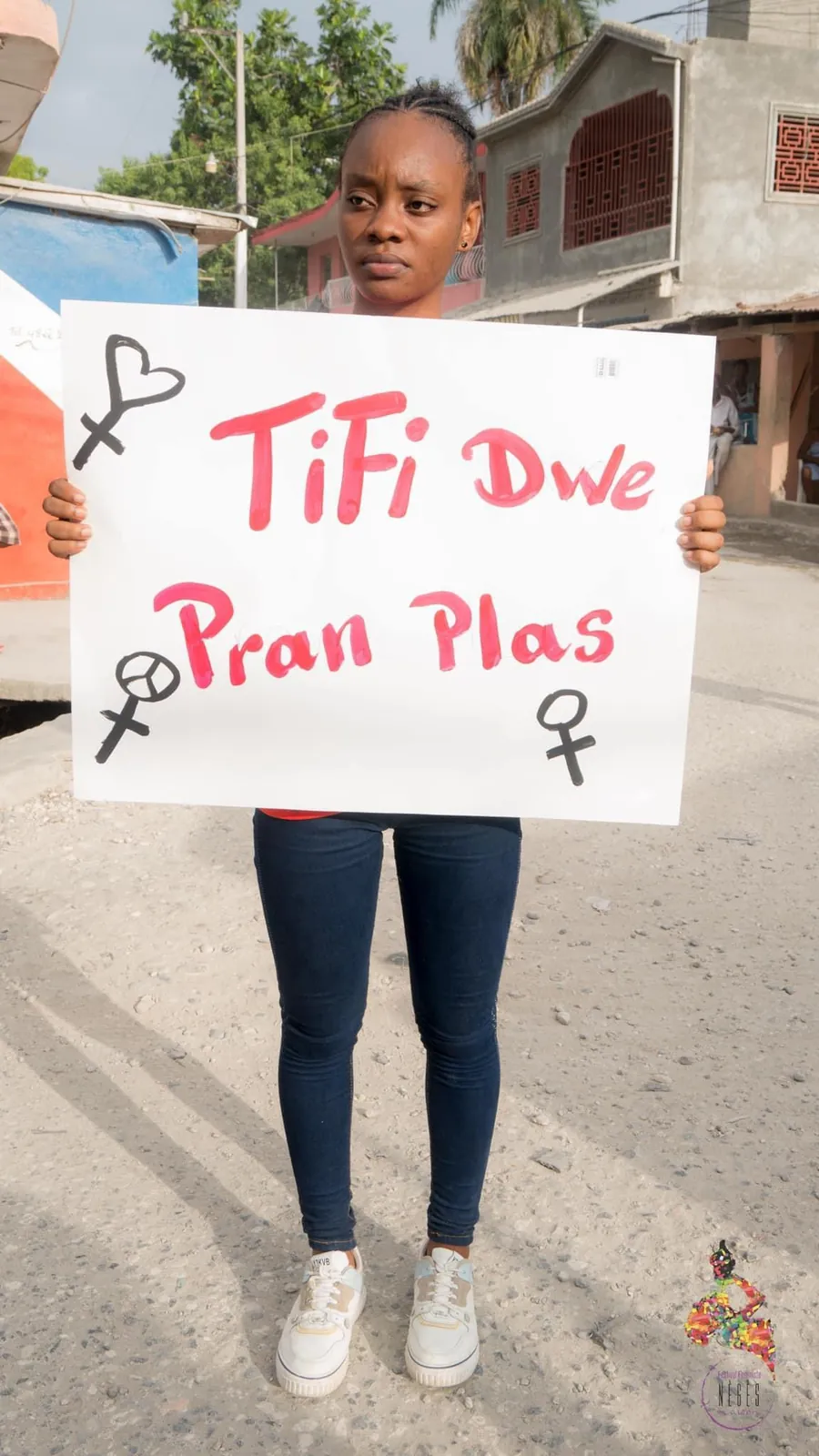 A young woman holding a placard that reads “TiFi Dwe Pran Plas”